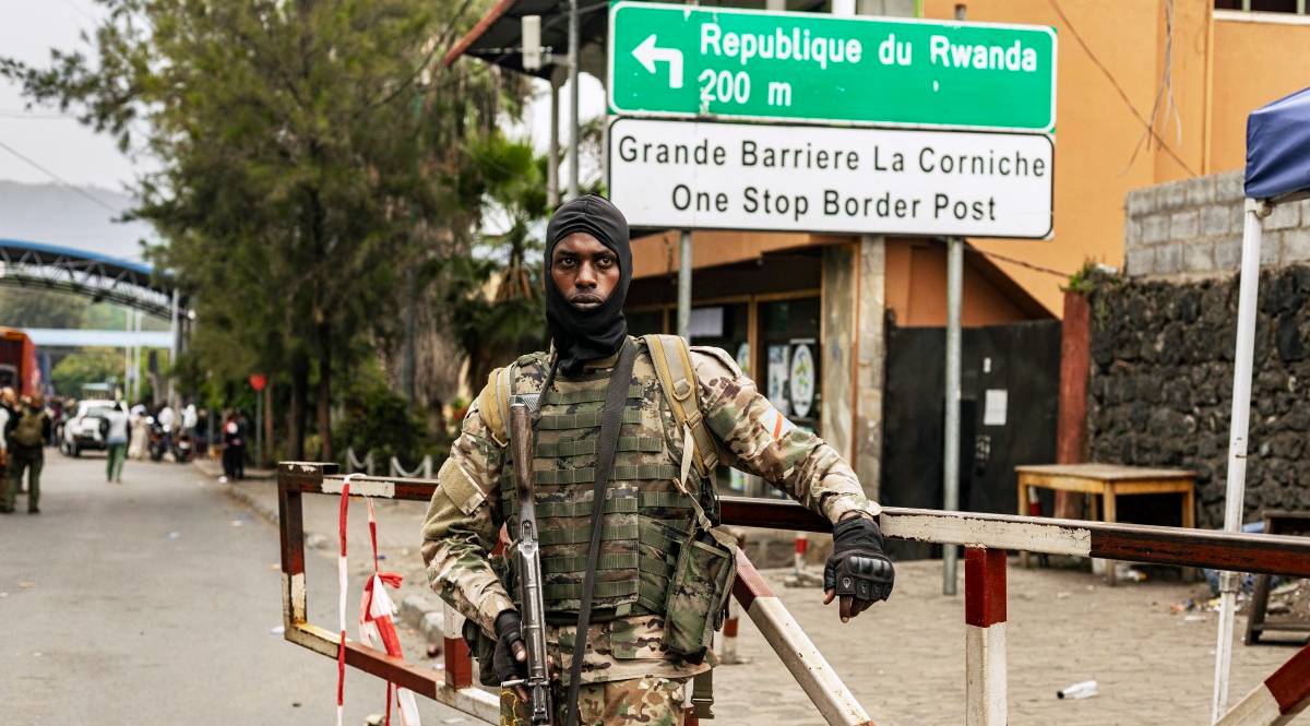 A member of the M23 rebel group stands at a road barrier, with a sign behind him pointing to the Rwanda border.