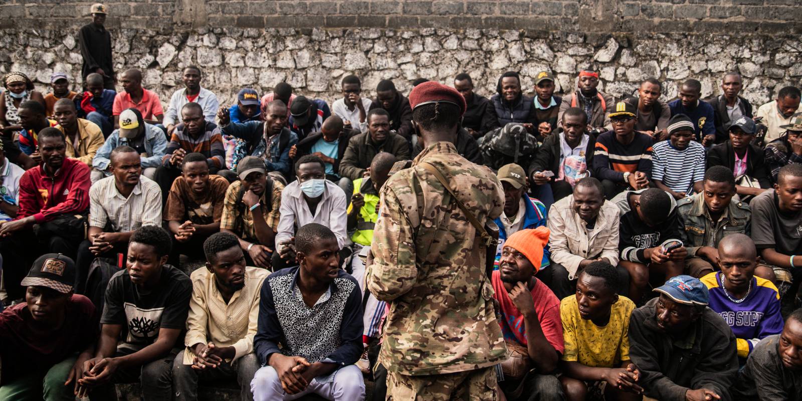 Dozens of Congolese sit in rows in front of a stone wall while a uniformed member of M23 collects their phones.