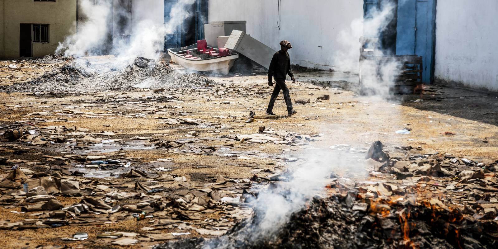 A man walks past several burning and smoking piles of materials outside a warehouse