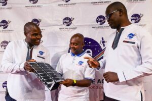 Before a KSA backdrop two Kenyan engineers hold a small satellite while a third points to it and speaks.