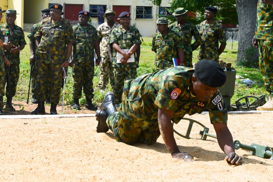 Nigerian IED training A soldier crawls in a training area with a metal detector while soldiers look on from the background.