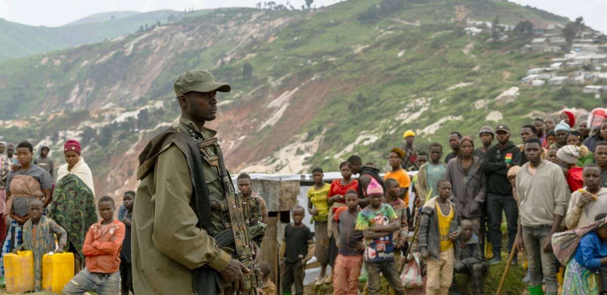 M23 soldier at coltan mining pits in Rubaya, eastern DRC A soldier stands amid several dozen local residents with a partially mined hill looms behind them.
