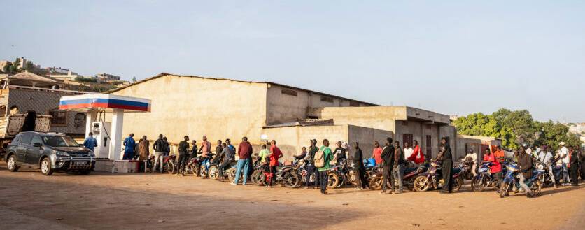 A line of motorcyclists starting at a petrol pump wraps around a building and down the street.