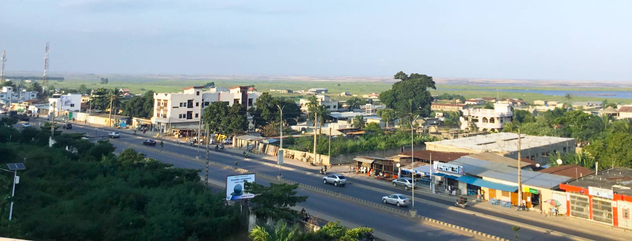 Side-aerial view of the city, with trees and the highway in the foreground and the green land beyond extending to the horizon.