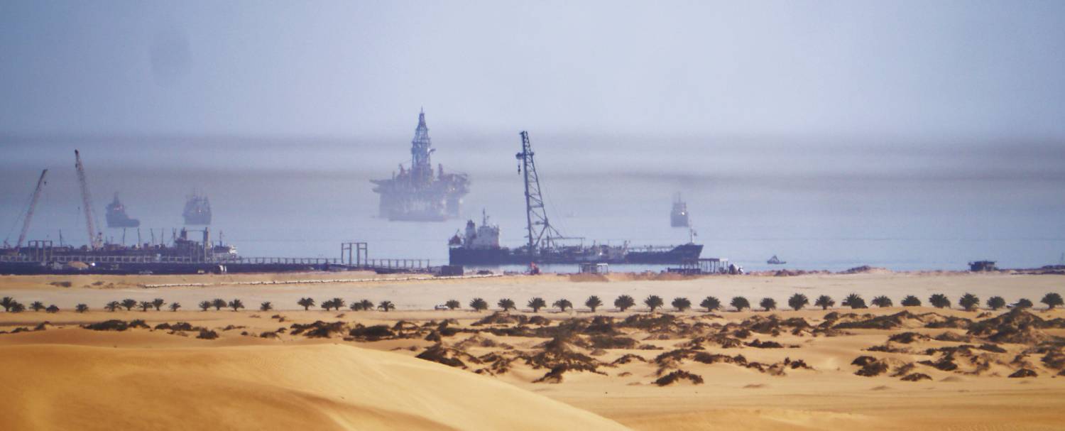 View of Walvis Bay, Namibia, with Dune 7 in the foreground, port infrastructure on the shore, and an oil platform in the Bay.